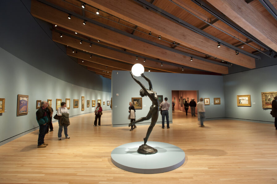Inside the Crystal Bridges museum. A sculpture of a woman holding a glowing ball is in the center of the room. Paintings line the walls.
